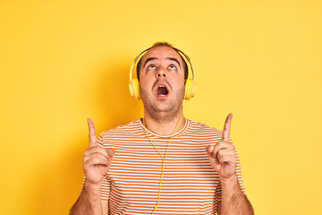 Poster - Young man listening to music using headphones standing over isolated yellow background amazed and surprised looking up and pointing with fingers and raised arms.