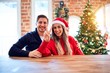 © Krakenimages.com - Young couple wearing santa claus hat sitting on chair and table around christmas tree at home showing and pointing up with fingers number two while smiling confident and happy.