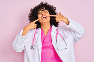 Sticker - Young arab doctor woman with curly hair wearing stethoscope over isolated pink background smiling cheerful showing and pointing with fingers teeth and mouth. Dental health concept.