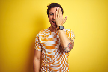 Wall Mural - Young handsome man with tattoo wearing striped t-shirt over isolated yellow background covering one eye with hand, confident smile on face and surprise emotion.