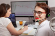 © Ihor - Cheerful young female customer support agent smiling to the camera, her colleague working on background