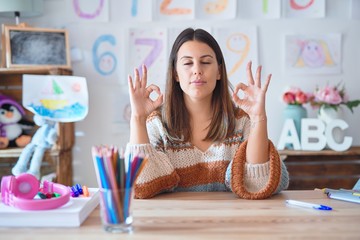 Canvas Print - Young beautiful teacher woman wearing sweater and glasses sitting on desk at kindergarten relax and smiling with eyes closed doing meditation gesture with fingers. Yoga concept.