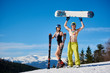 Smiling romantic couple, sexy skier woman in swimsuit, helmet and boots and man with bare torso holding snowboard standing on snowy hill on copy space background of blue sky and winter mountains.