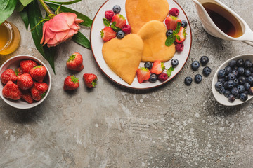 Wall Mural - top view of heart shaped pancakes with berries on grey concrete surface with blooming rose, honey and maple syrup