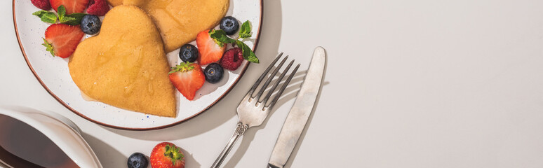 Wall Mural - top view of delicious heart shaped pancakes with berries near cutlery and maple syrup on white background, panoramic shot