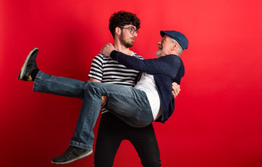 Wall Mural - Young man holding senior father in a studio on red background.