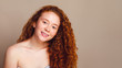 © Shauerman - beautiful girl with thick red long curly hair smiles in the studio on a beige background. Copy space