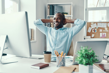 Sticker - Handsome young African man in shirt keeping hands behind head and smiling while working in the office