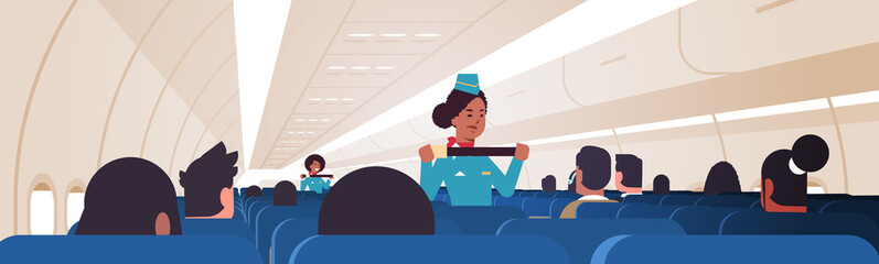 Wall Mural - stewardess explaining passengers how to use seat belt fastening in emergency situation african american flight attendants in uniform safety demonstration concept airplane board interior horizontal