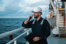 Marine Deck Officer Or Chief Mate On Deck Of Offshore Vessel Or Ship , Wearing PPE Personal Protective Equipment - Helmet, Coverall. He Holds VHF Walkie-talkie Radio In Hands.