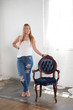 Beautiful young blonde woman wearing white tank top and blue jeans stands next to vintage blue upholstered chair