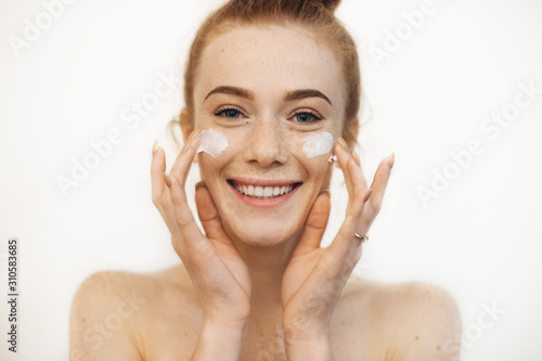 Fotografía Portrait of a charming young woman looking at camera laughing while playing with both hands cream on her cheeks