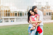 © Look! - Shapely girl in blue jeans in wristwatch huggs with husband near lawn in front of building. Happy young man with red cap on pants posing with attractive woman.