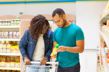 Wall Mural - Focused people looking at bottle while choosing goods. Young couple shopping together. Shopping concept