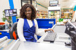 Happy positive female cashier working in grocery store. Young African American woman wearing uniform, posing at cash register counter in supermarket. Cashier concept