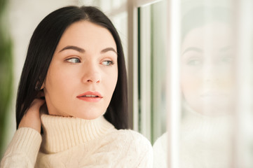 Portrait of a brunette girl at the window with reflection closeup. Young woman in a white knitted dress on a background of a large window and copy space.