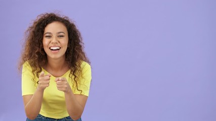 Sticker - Attractive happy african young woman in yellow t-shirt pointing to the camera and then advertising to the at copy space while looking at the camera over purple background isolated 