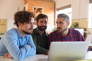 Male coworkers using laptop together, watching content and talking. Business colleagues in casual meeting in contemporary office space. Colleague discussion concept