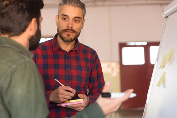 Wall Mural - Male coworkers standing at flipchart, discussing project. Business colleagues in casual working together in contemporary office space. Training concept