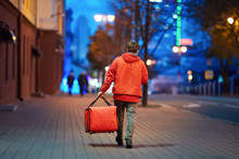 Young Deliveryboy Walking With Red Thermal Bag On Night City Street. Man Of Delivery Service In Hurry To Deliver An Order. Delivery Service Goes To Give The Order Quickly To The Client At Night