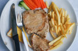 A pork steak consisting of french fries, carrots, knives and grilled pork on a white plate