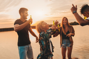 Group of young friends having fun drinking beer and dancing on pier by the lake.