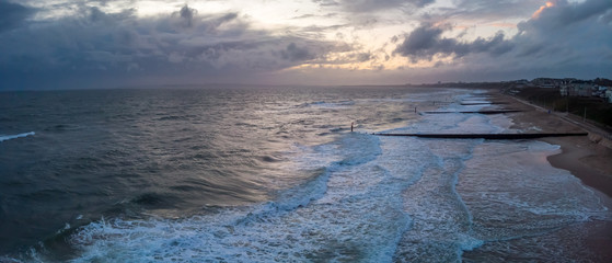 An aerial panoramic view of a choppy sea with crashing waves, groynes (breakwater), sandy beach and city in the background under a stormy cloudy grey sky