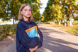 © byswat - Serious teenager girl with smart look, stands summer park, background autumn trees, free space copy text. In hands notebook textbooks and notes, preparation school college. Warm clothes sweater.
