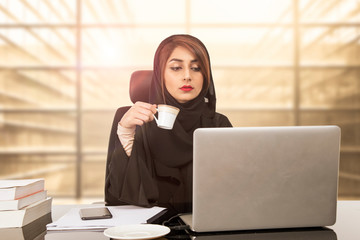 Arab Young business woman sitting at her desk in an office, working on a laptop computer and drinking coffee or tea.