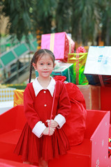 Wall Mural - Cheerful Asian little girl in red dress carrying a red big bag in winter season and happy new year festival against christmas background.