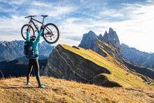 Young Man Raising Mountain Bike To The Sky On Seceda Mountain Peak At Sunrise. Puez Odle, Trentino, Dolomites, Italy.