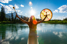 A Backlit And Rear View Of A Spiritual Woman Holding A Powwow Drum During A Mindful Ritual In Nature, Bright Sun Gives Atmospheric Mystical Lens Flare