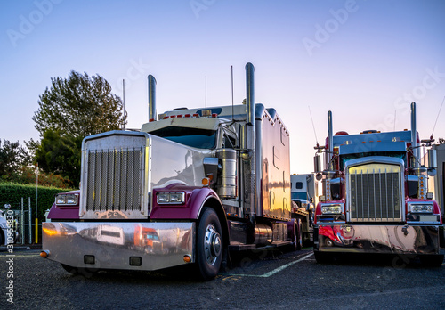 Big rigs classic bonnet semi trucks standing in row on truck stop parking lot at evening time reflecting sunset and all around by lot of chrome parts