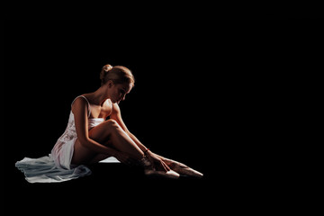 Graceful ballet or gymnast sitting on a black background wears shoes - pointe shoes. dancer in a white dress. Isolated. Dancer posing in Studio with copy space. black background    