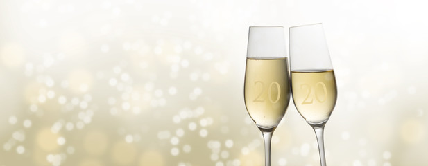 Happy New Year 2020, two champagne glass flutes toasting against a light golden bubble bokeh background, party concept in panoramic format with copy space