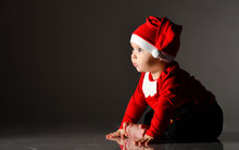 Fascinated Barefooted Infant Child Boy Toddler Is Sitting In Santa Claus Costume On Ice On Dark Looking At Copy Space