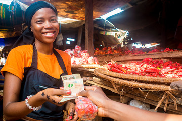 Wall Mural - young african woman selling tomatoes in a local african market collecting money from a paying customer