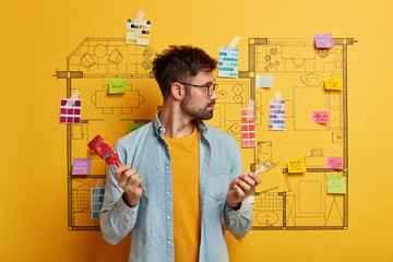 Making repairs. Serious male painter stands with two paintbrushes, looks attentively at room sketch, busy improving house, wears transparent glasses, involved in building, redecorates apartment