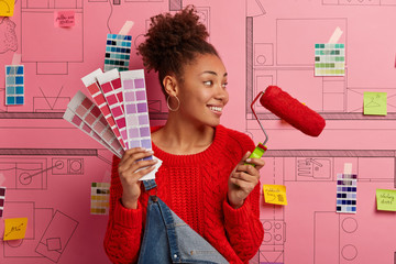 Pleased female painter with curly combed hair looks aside, notices something nice, stands with color samples, holds paint roller, wears red sweater, busy with painting and redecoration of house