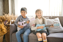 Happy Elementary Age Asian Little Kid Is Smiling  While Playing A Ukulele During A Private Music Learning Lesson At Home