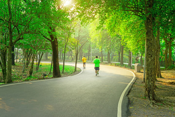 A black asfalt concrete jogging track in a public park, two people wearing yellow and green T shirt running on curve shape way under evergreen leaves trees and sunshine morning