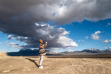 A Young Woman In A Native American Patterned Vintage Jacket And Mustard Hat Explores Sand Dunes In California Under Approaching Storm Clouds
