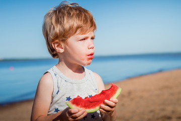 little cute redhead boy in grey shirt holding slice of watermelon with dripping juice and got smeared face with it at the sunny summer beach