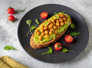 toast with avocado, roasted chickpeas, cherry tomatoes, herbs on a dark plate. healthy vegan food. gray concrete background, side view