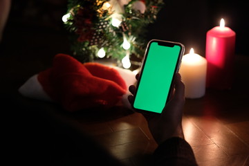 Wall Mural - over shoulder of one hand holding green screen smartphone. Blur christmas tree, hat and candle lights. black background