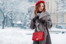 Happy Smiling Woman Enjoying Winter Holidays, Posing In Snow Covered Street Of European City. Model Wearing Beret, Grey Scarf, Coat, Gloves, With Red Quilted Bag. Copy, Empty Space For Text 