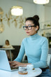 © pressmaster - Young serious woman with wireless earphones sitting in front of laptop by table