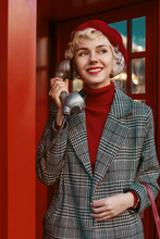 Fashionable Happy Smiling Blonde Woman Wearing Red Beret, Turtleneck, Checkered Coat, Posing In Red Retro Phone Booth