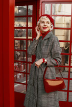 Fashionable Happy Smiling Blonde Woman Wearing Red Beret, Turtleneck, Checkered Coat, White Wrist Watch, Holding Stylish Leather Handbag,  Posing In Red Retro Street Phone Booth. 