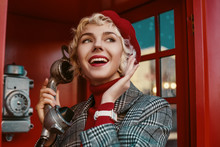 Fashionable Happy Smiling Blonde Woman Wearing Red Beret, Turtleneck, Checkered Coat, White Wrist Watch, Talking On The Retro Phone, Posing In Red  Call Box. Copy, Empty Space For Text